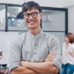 Portrait of successful handsome executive businessman smart casual wear looking at camera and smiling, arms crossed in modern office workplace. Young Asia guy standing in contemporary meeting room.